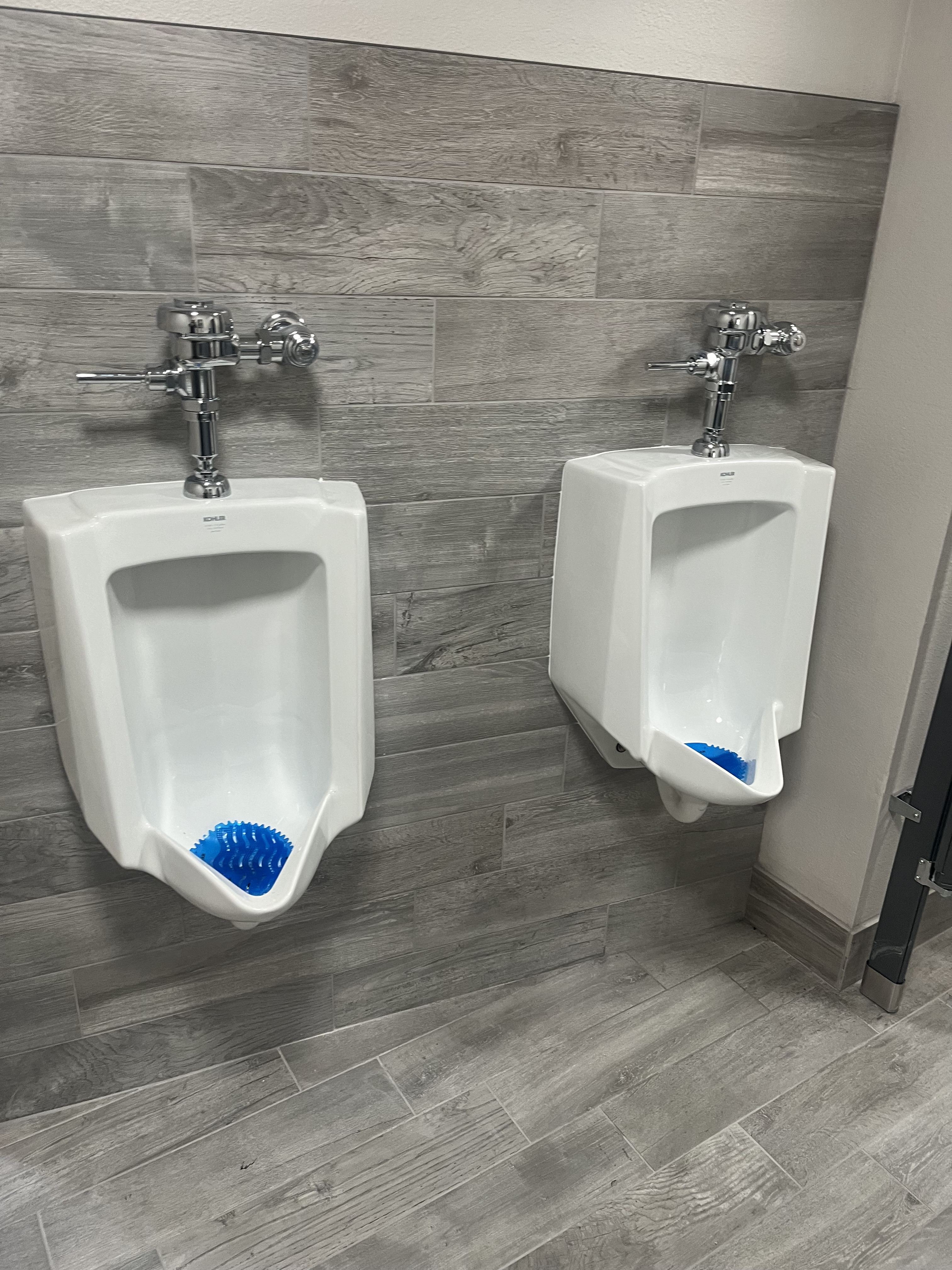 Two white urinals with blue urinal screens are mounted on a wall covered with gray wood-look tiles in a public restroom.