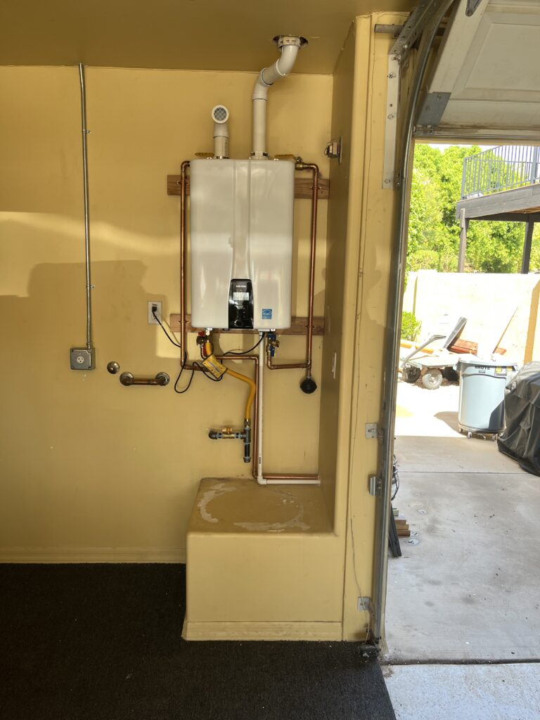 A white, rectangular tankless water heater is mounted on a yellow wall, with copper and PVC pipes connecting to it, in what appears to be a garage or utility space.