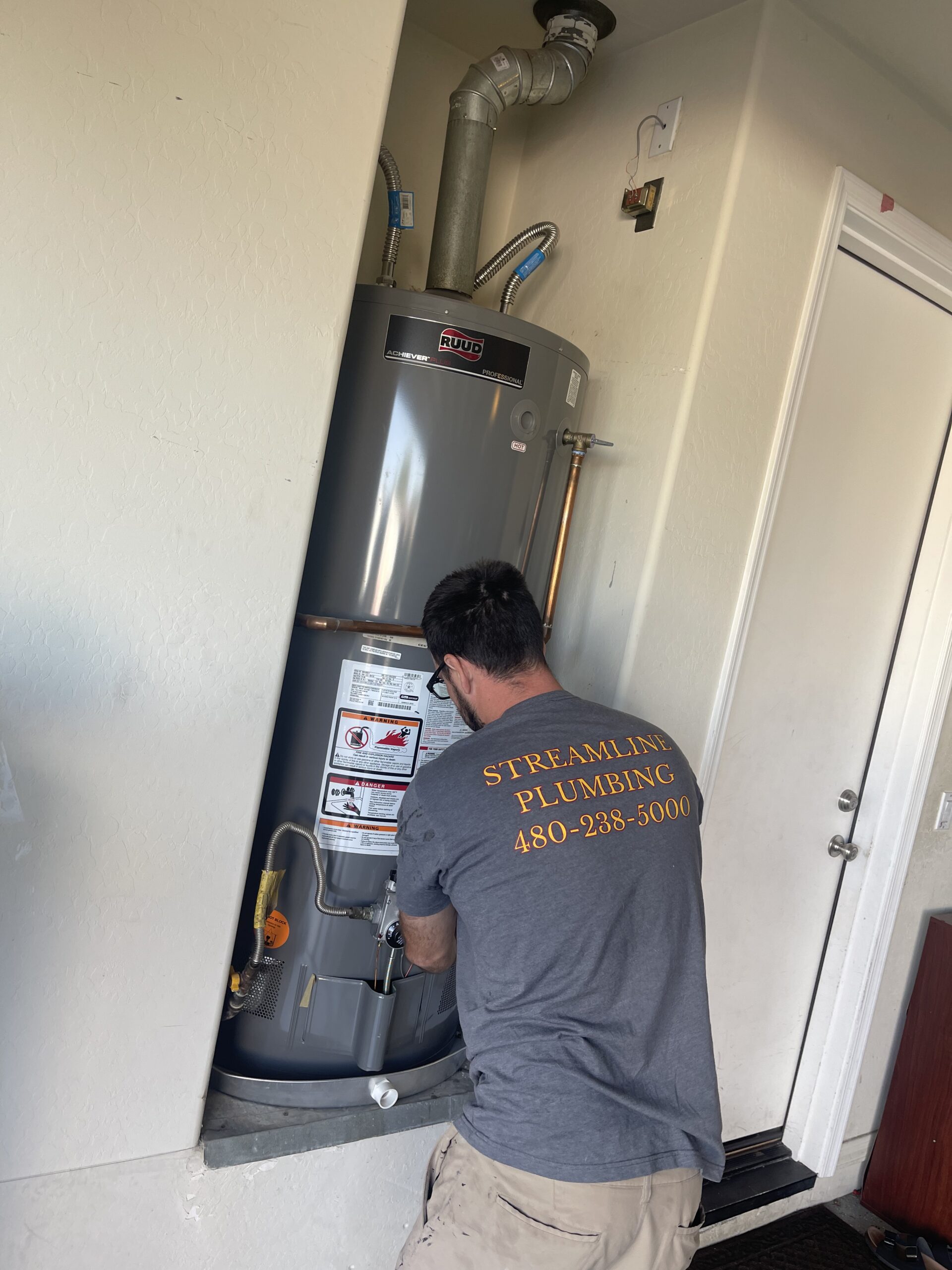 A plumber, wearing a "Streamline Plumbing" t-shirt, kneels to work on the lower section of a gray Ruud water heater in a narrow utility space.