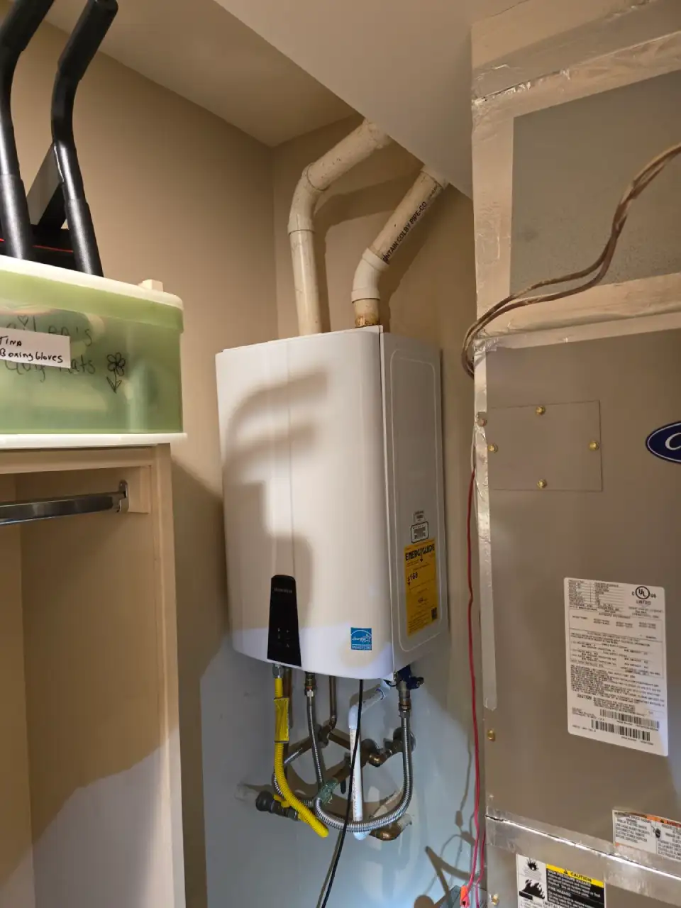 A compact, white, tankless water heater is mounted on a wall, with pipes connecting to it from the top and bottom, next to a large HVAC unit.