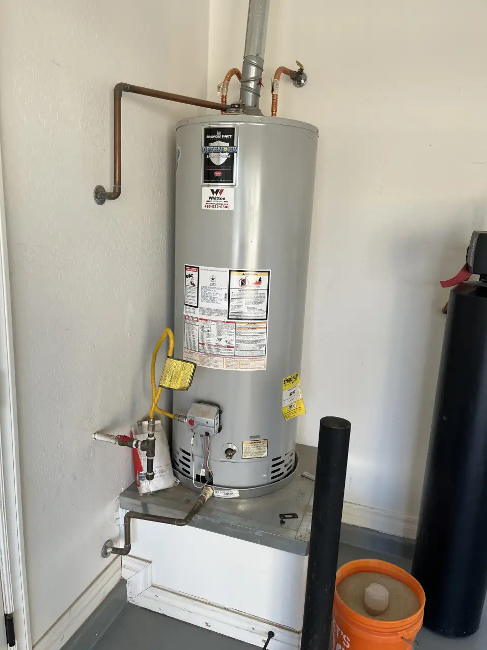 A tall, light gray Bradford White water heater sits on a raised platform, with visible plumbing and a nearby water softener unit.