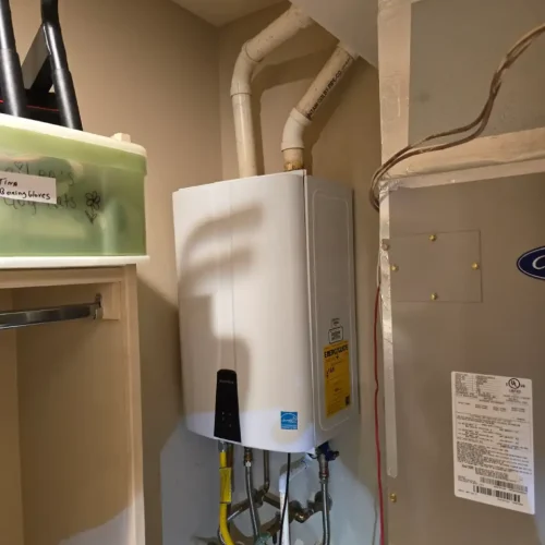 A compact, white, tankless water heater is mounted on a wall, with pipes connecting to it from the top and bottom, next to a large HVAC unit.